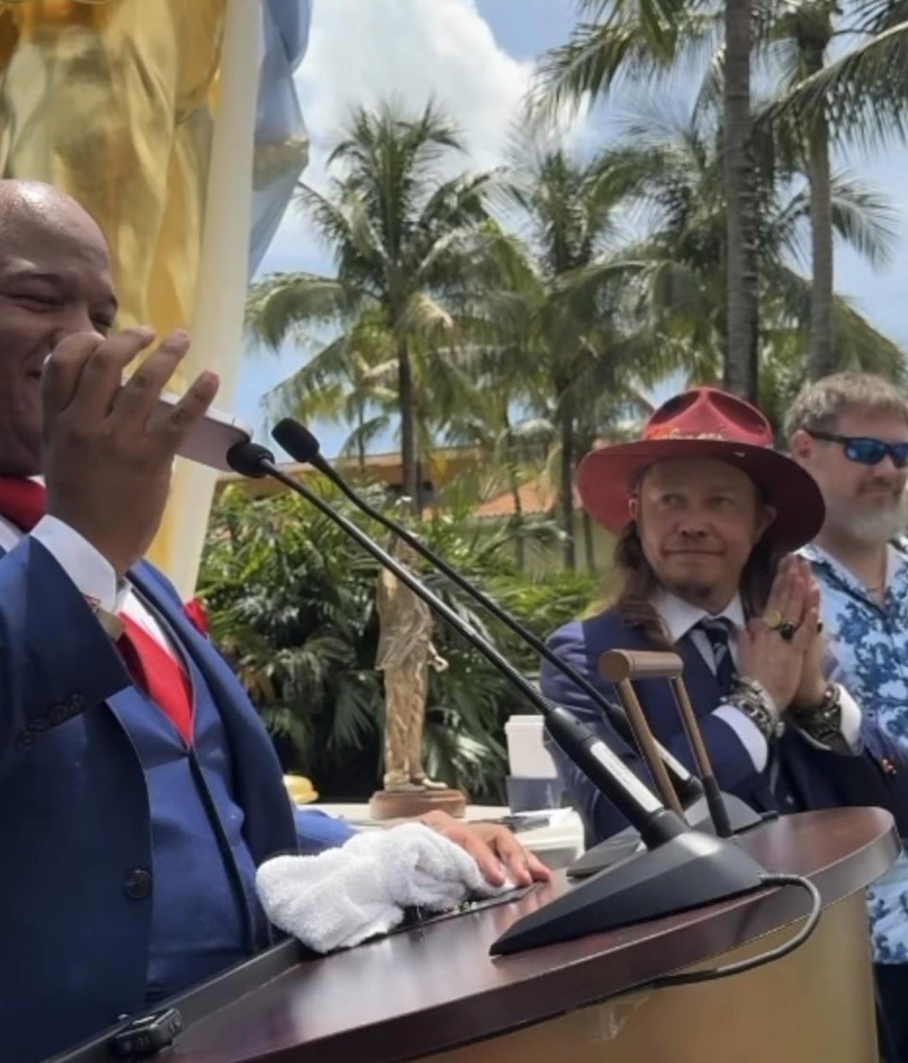 Brock Pierce in a red hat applauding at the Don Colossus statue unveiling, beside a speaker at a podium