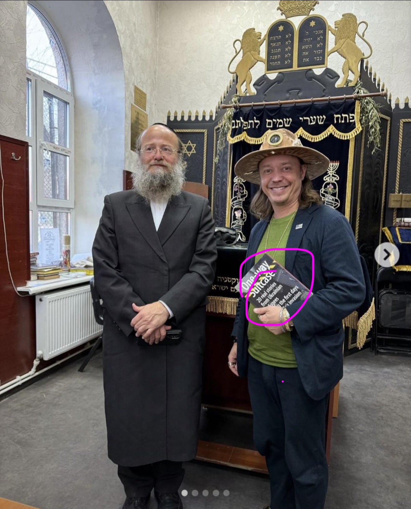 Brock Pierce standing with a rabbi in a synagogue, holding a book titled One Way Suitcase